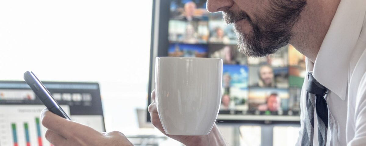 Businessman drinking coffee while reading cell phone screen with video chat computer and data charts