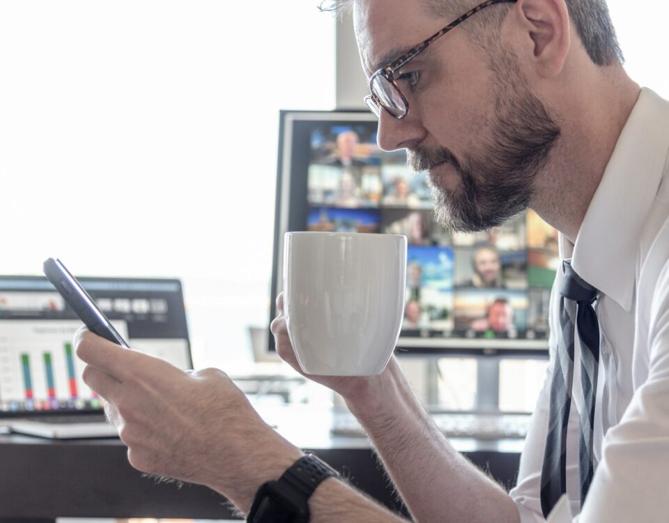 Businessman drinking coffee while reading cell phone screen with video chat computer and data charts